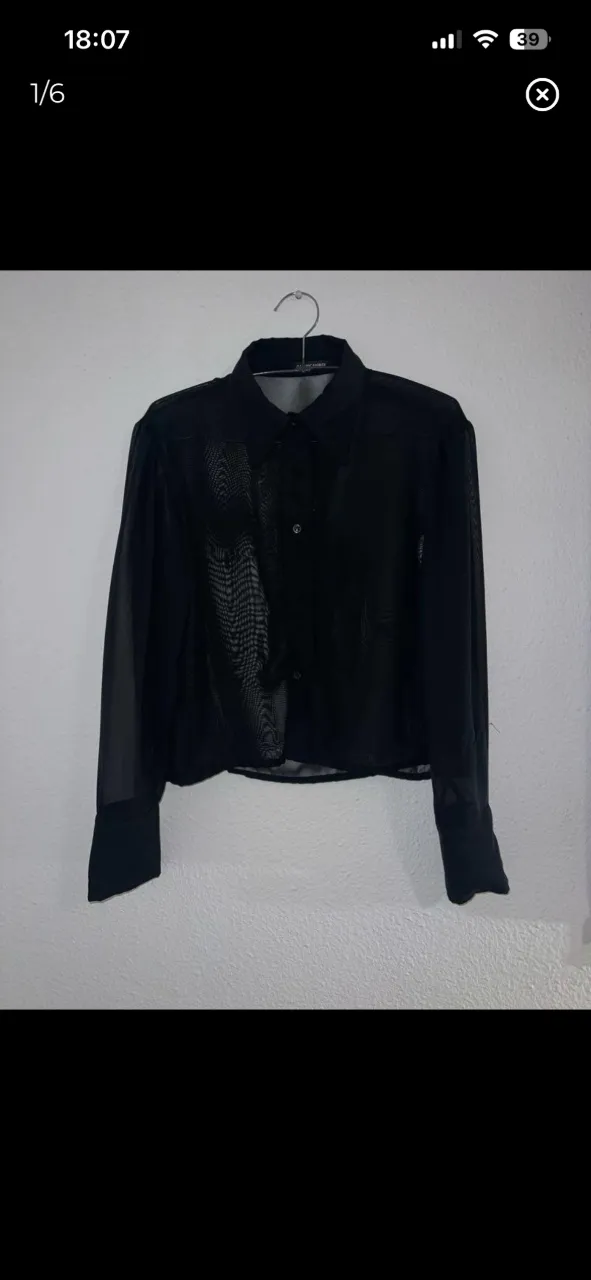 Camisa negra de manga larga, con cuello y botones al frente. La tela es transparente tipo de tul. Es corta. 