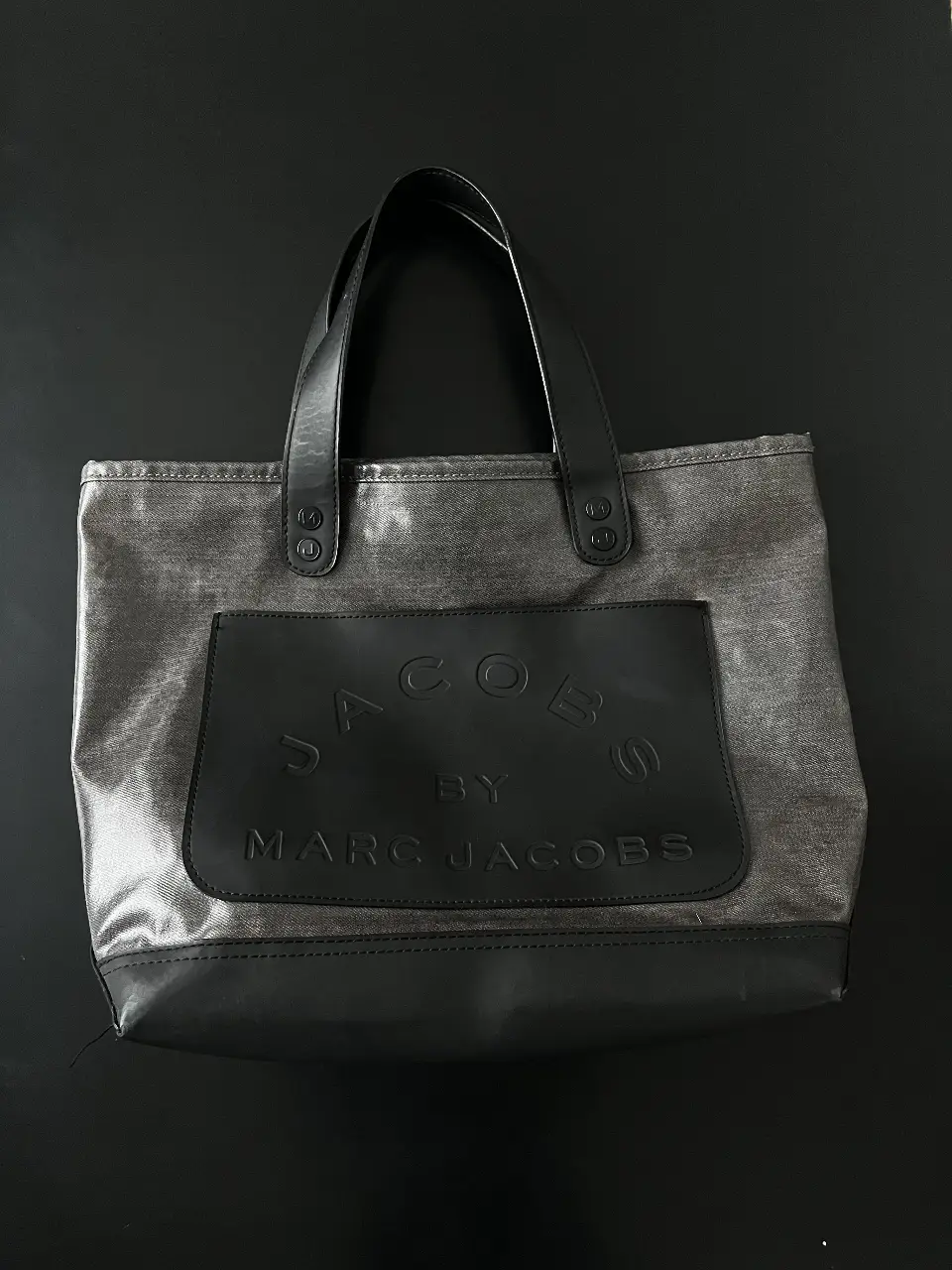 Bolso tote de la marca Marc Jacobs, confeccionado en lona gris con detalles en cuero negro. Cuenta con un bolsillo frontal con el logo de la marca y asas de cuero. Ideal para llevar tus pertenencias con estilo y comodidad.