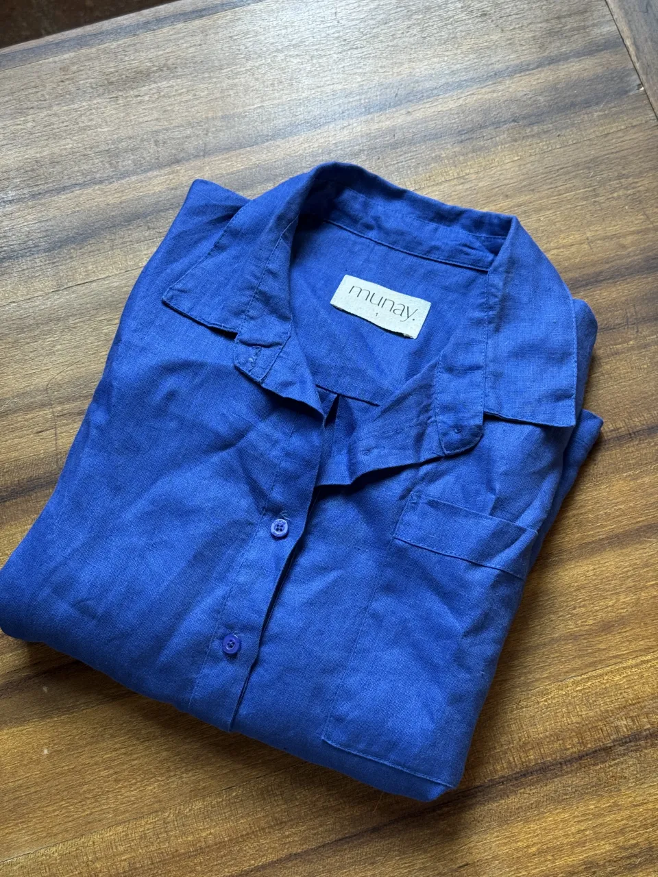 Camisa de lino azul vibrante, con cuello clásico y botones al frente. Posee dos bolsillos en el pecho y dos bolsillos inferiores amplios. Un detalle de etiqueta naranja con el logo de la marca le da un toque distintivo. Ideal para un look casual y fresco.