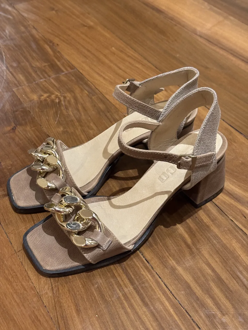 Sandalias de mujer color marrón con detalles dorados. Diseño elegante con cadena en la parte delantera y ajuste de hebilla en el tobillo. Ideales para un look sofisticado.