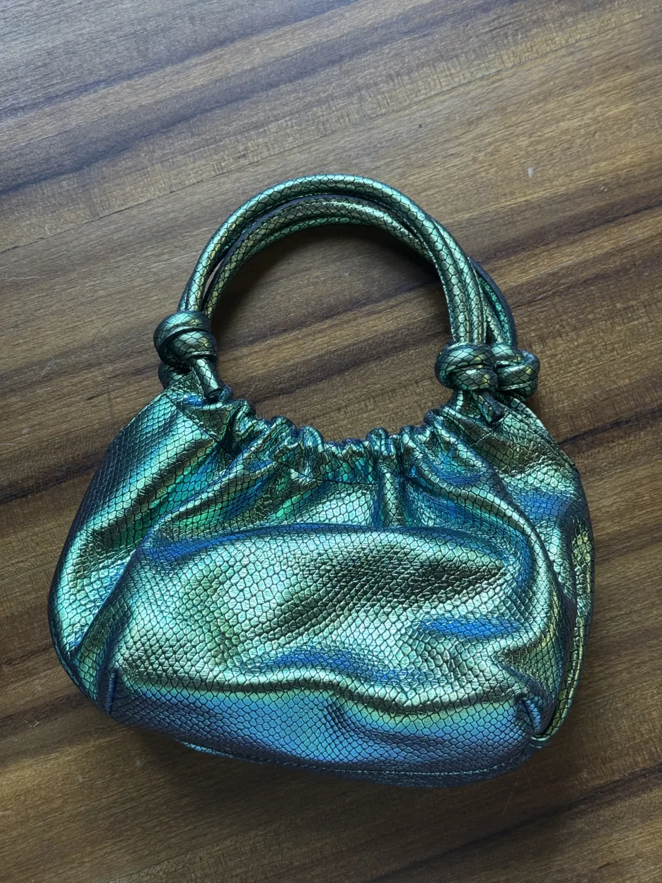 Cartera súper canchera con efecto tornasolado y textura de reptil. Tiene un cierre interno y cierre principal con imán. Las manijas son anudadas y le dan un toque único. ¡Ideal para darle un toque de brillo a cualquier look!