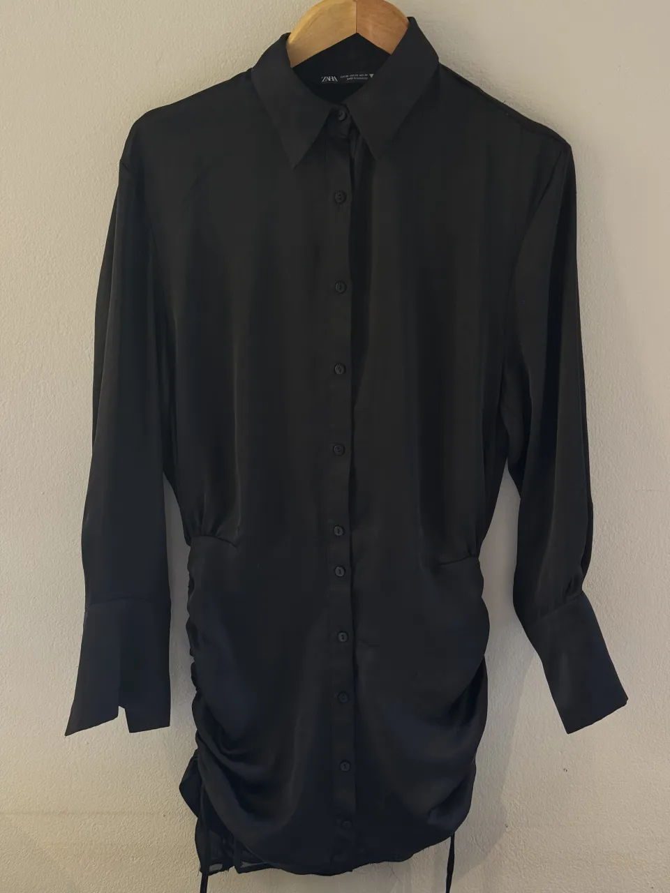 Camisa larga negra con fruncido ajustable en el lateral. Mangas largas con puños abotonados y cuello de camisa. Ideal para una salida elegante o una fiesta. Tiene un pequeño desgaste en los hombros