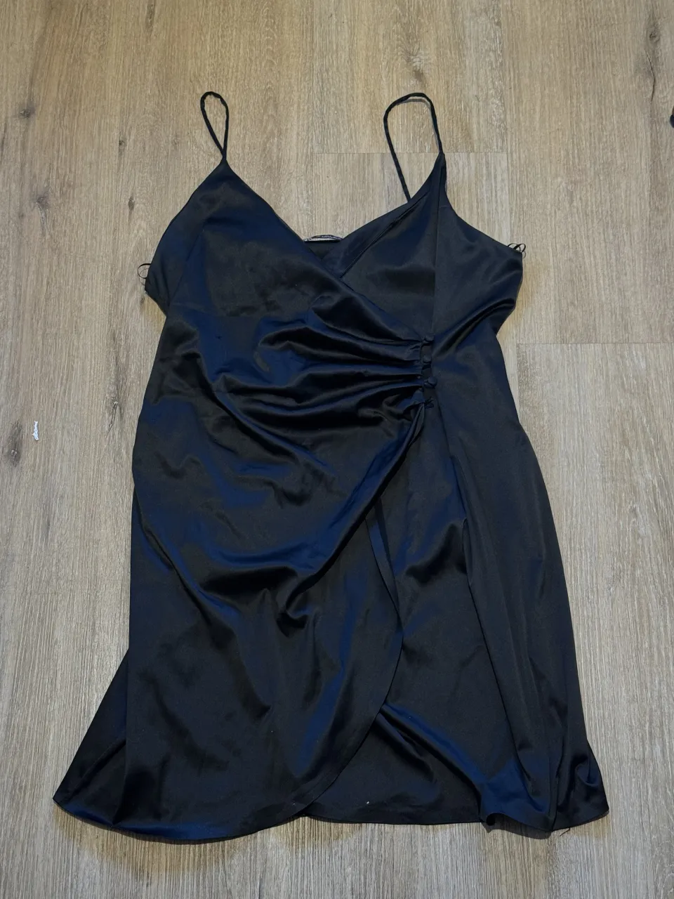 Vestido negro corto con tiras finas y detalle de fruncido y botones en un lateral. Posee un corte cruzado en la falda. DETALLES: le falta un boton y tiene un hilito salido en la parte izquierda arriba adelante (tercera foto)