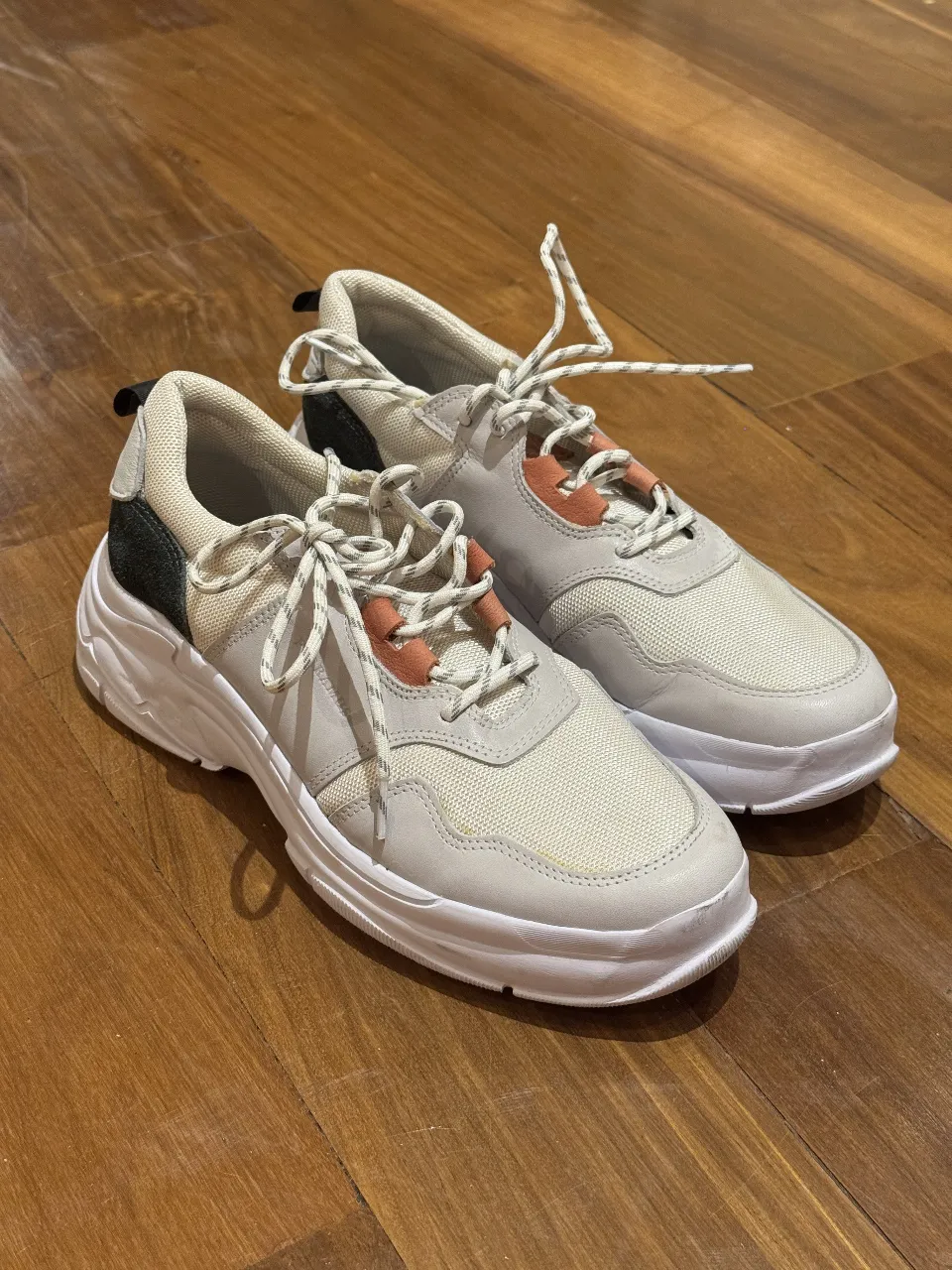 Zapatillas deportivas blancas con detalles en negro y beige. Suela gruesa y cordones a tono. Ideales para un look casual y cómodo.