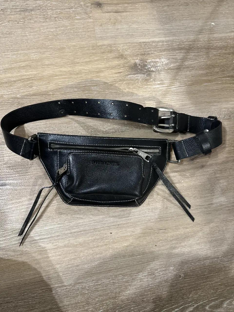 Riñonera de cuero negro con detalles de costura en blanco. Cuenta con un bolsillo frontal y otro principal con cierre. La correa es ajustable y tiene hebilla plateada. Puede ir al hombro o cintura.