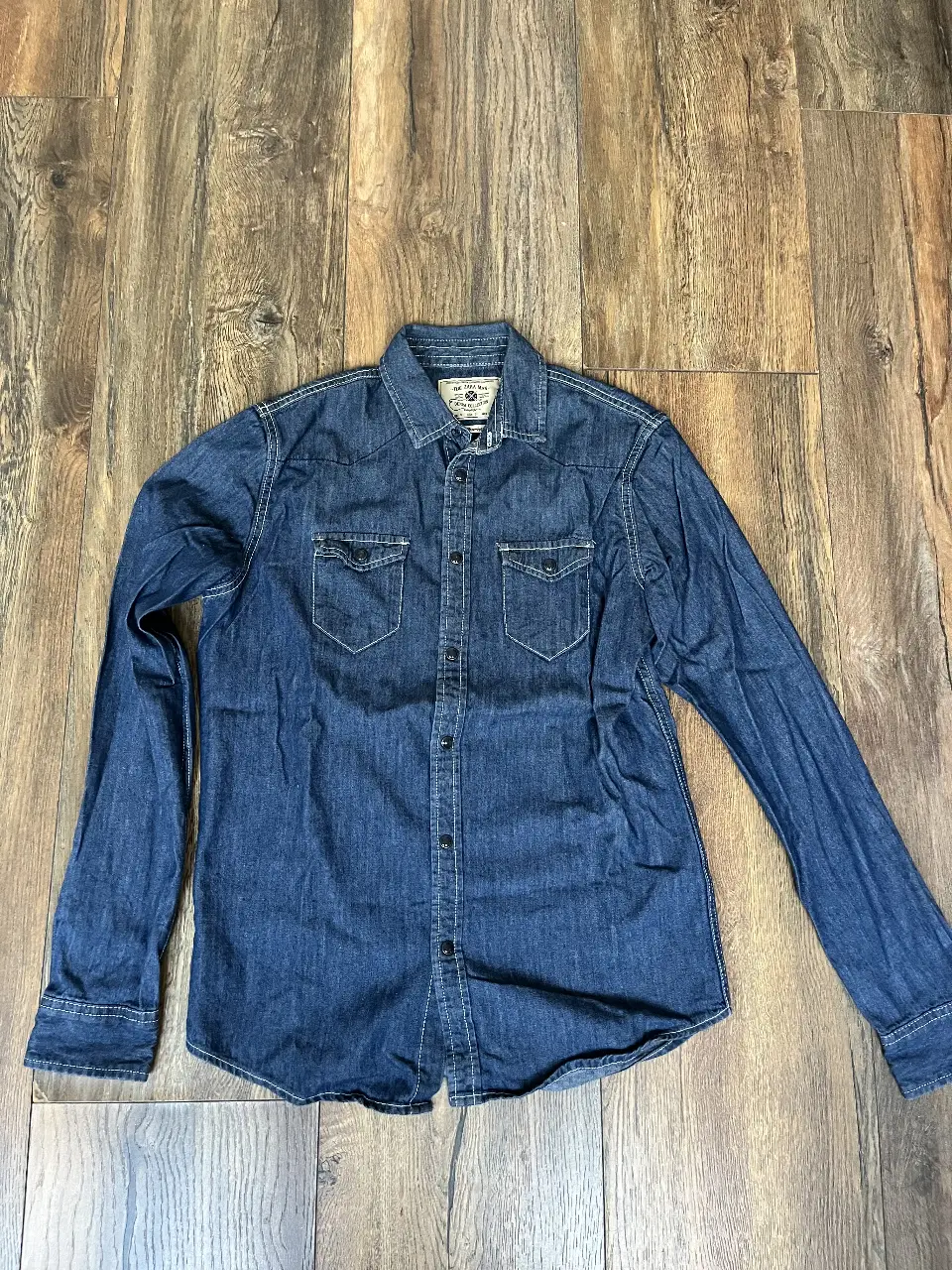 Camisa de jean azul oscuro de hombre, manga larga, con dos bolsillos delanteros y cierre con botones. Ideal para un look casual y versátil.