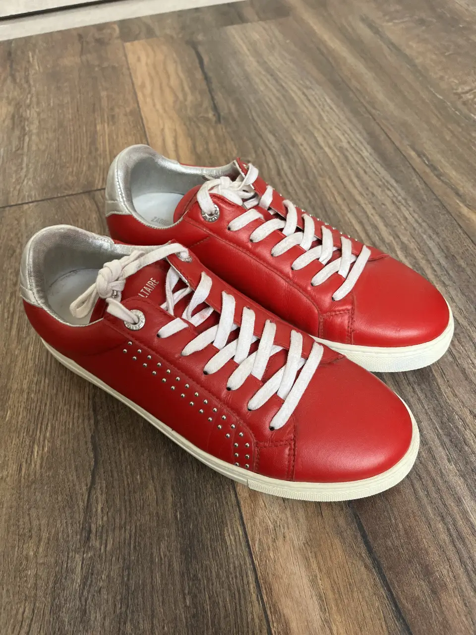 Zapatillas rojas de cuero con detalles plateados y tachas. Tienen cordones blancos y suela blanca. Ideales para un look casual y moderno.