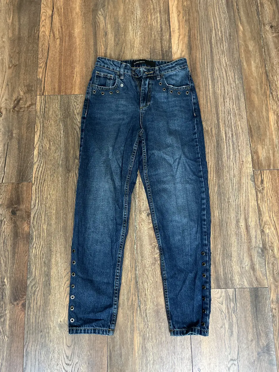 Jeans de mujer, tiro alto, color azul. Estolo “mom”. Con detalles de arandelas en los bolsillos delanteros y en los laterales de los tobillos. Un clásico que no puede faltar en tu guardarropa. Talle 0. 