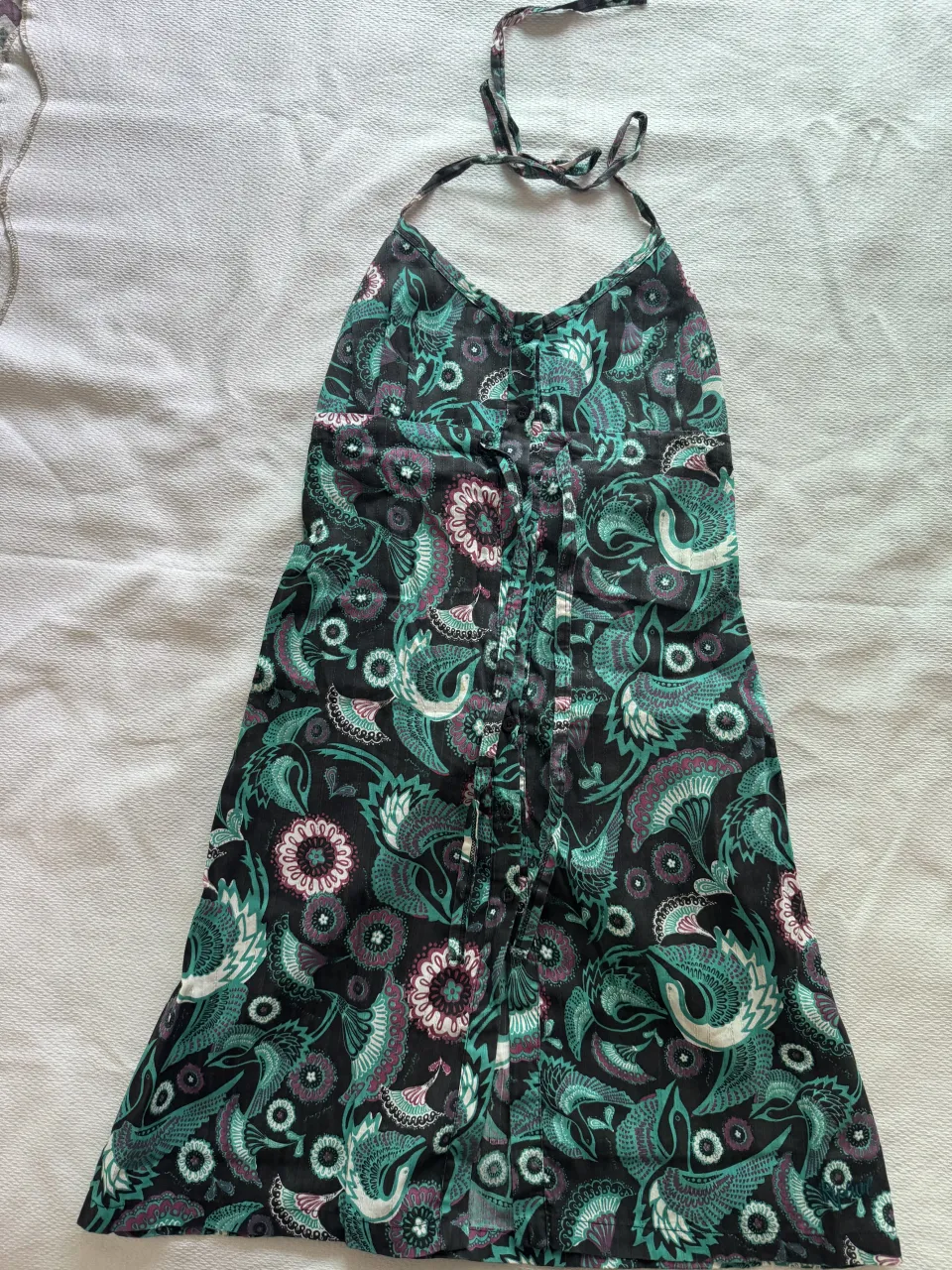 Vestido corto con estampado floral y de aves en tonos verde, rosa y negro. Posee tiras finas que se anudan en el cuello y botones en la parte delantera.