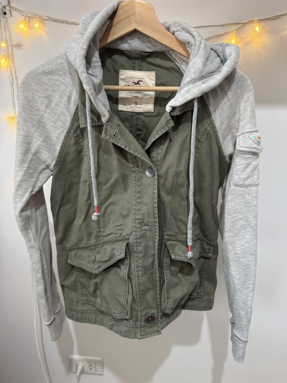 Campera verde con mangas grises y capucha. Tiene bolsillos frontales y cordón ajustable en la cintura.