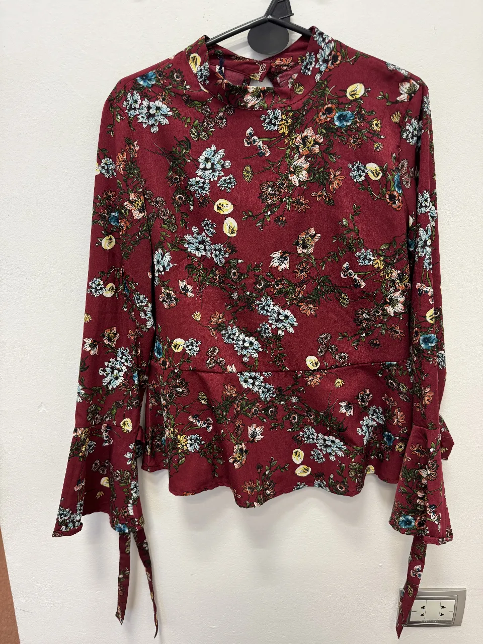 Blusa de mujer con estampado floral en tonos rojos y detalles en otros colores. Cuello alto y mangas largas con lazos en los puños. Ideal para un look elegante y femenino.