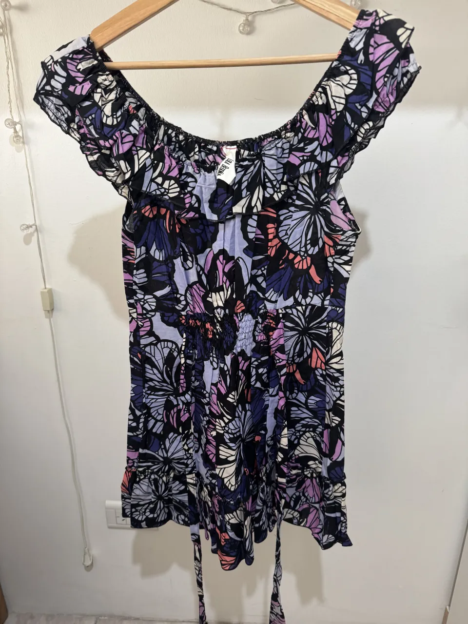 Vestido corto estampado con flores y mariposas en tonos violeta, azul, negro, blanco y coral. Escote con volados y elástico. Cintura con elástico y lazo para atar. Mangas cortas con volados.