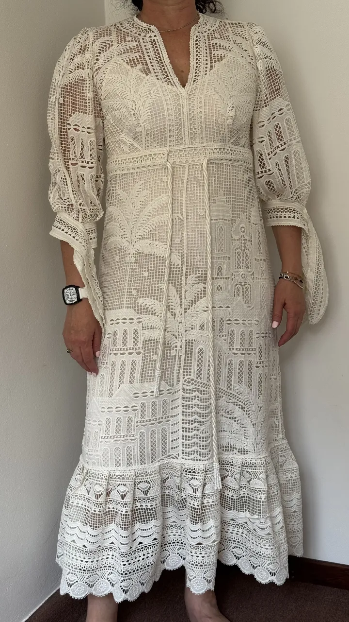 Vestido blanco largo de encaje con detalles intrincados y mangas acampanadas. Posee un cinturón de soga fino que marca la cintura y un ruedo con volados. Ideal para eventos de día o ceremonias.