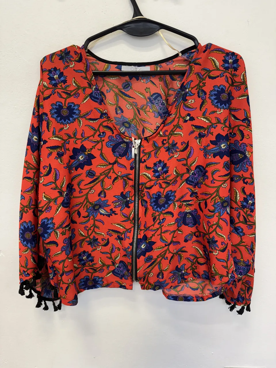 Blusa de mujer con estampado floral en tonos rojos y azules. Tiene cierre frontal, mangas largas con detalles de borlas y un corte holgado. Ideal para un look bohemio y casual.