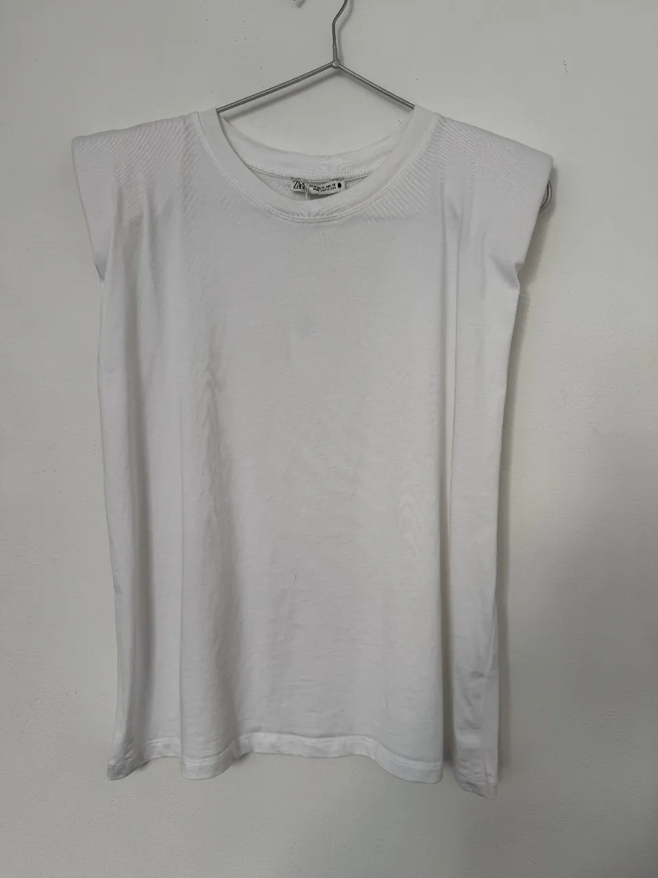 Remera blanca de mujer, con hombreras y cuello redondo. Marca Zara, talle M. Ideal para un look casual y moderno.