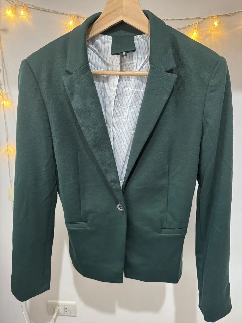 Blazer de color verde oscuro, con un botón al frente y bolsillos laterales. El interior está forrado con una tela a rayas finas blancas y negras.