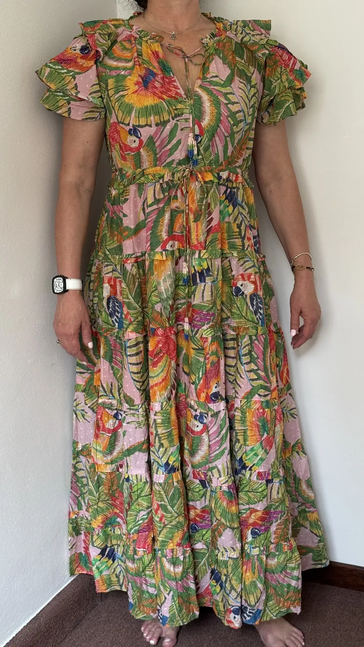 Vestido largo estampado con loros y hojas tropicales. Posee mangas cortas con volados, escote en V con cordones y detalle de canutillos. Cintura elastizada con lazo para ajustar. Falda con varios volados escalonados. Ideal para un look playero o de verano.