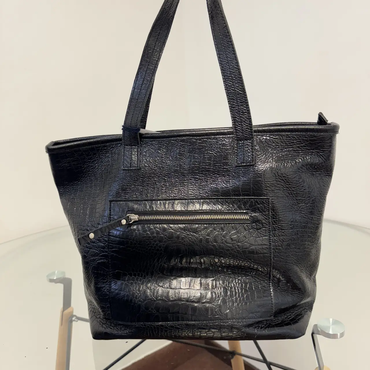 Cartera tote bag negra con textura símil cuero de cocodrilo. Posee un bolsillo frontal con cierre y manijas para llevar al hombro. Interior forrado en tela color rosa.