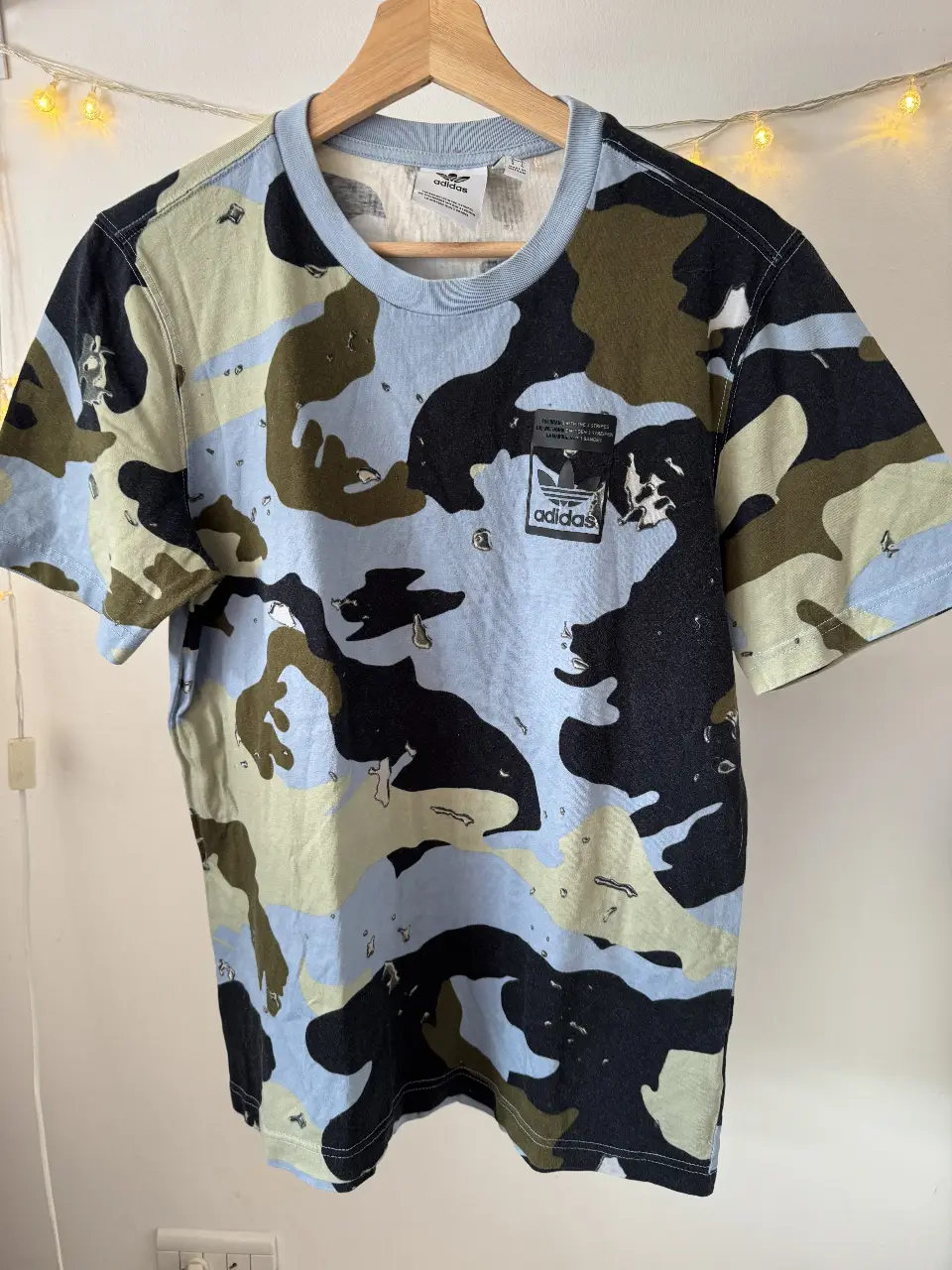 Remera Adidas con estampado de camuflaje en tonos azules, verdes y negros. Cuello redondo y logo de la marca en el pecho. Ideal para un look casual y urbano.
