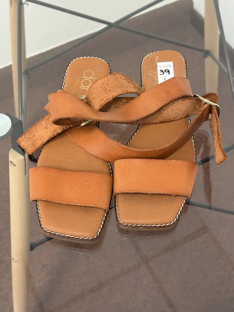 Sandalias planas de cuero marrón con detalles de costura en blanco. Diseño con tiras cruzadas y hebilla dorada. Ideales para un look casual y cómodo.