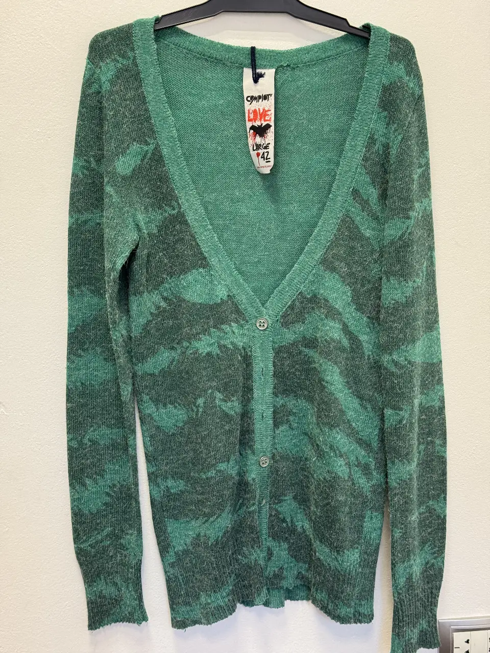 Cardigan de punto con diseño de camuflaje en tonos verdes y grises. Tiene cuello en V, botones y un estilo casual y cómodo. Ideal para un look relajado y moderno.