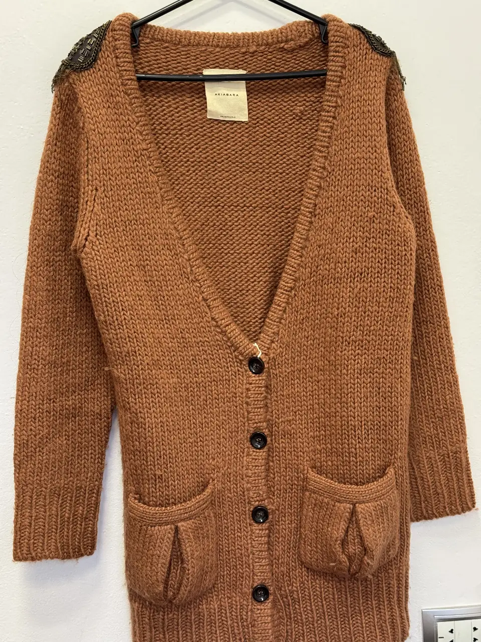Cardigan de tejido grueso color marrón con detalles de apliques en los hombros. Posee dos bolsillos delanteros y cierre con botones.