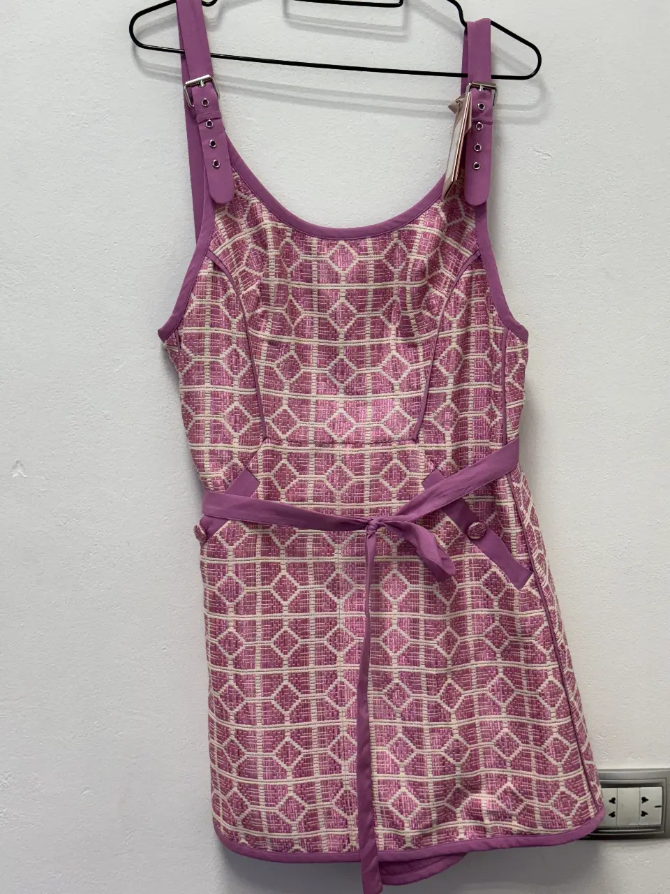 Vestido mini con estampado geométrico en tonos rosa y blanco. Diseño con tirantes ajustables, cinturón a juego y detalles de botones. Ideal para un look fresco y femenino.