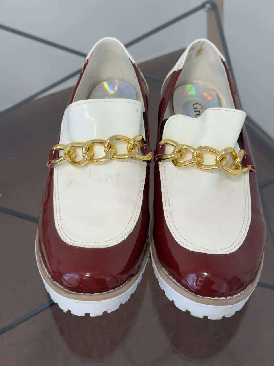 Elegantes mocasines de charol en borgoña y blanco, con detalle de cadena dorada. Ideales para un look sofisticado y moderno.