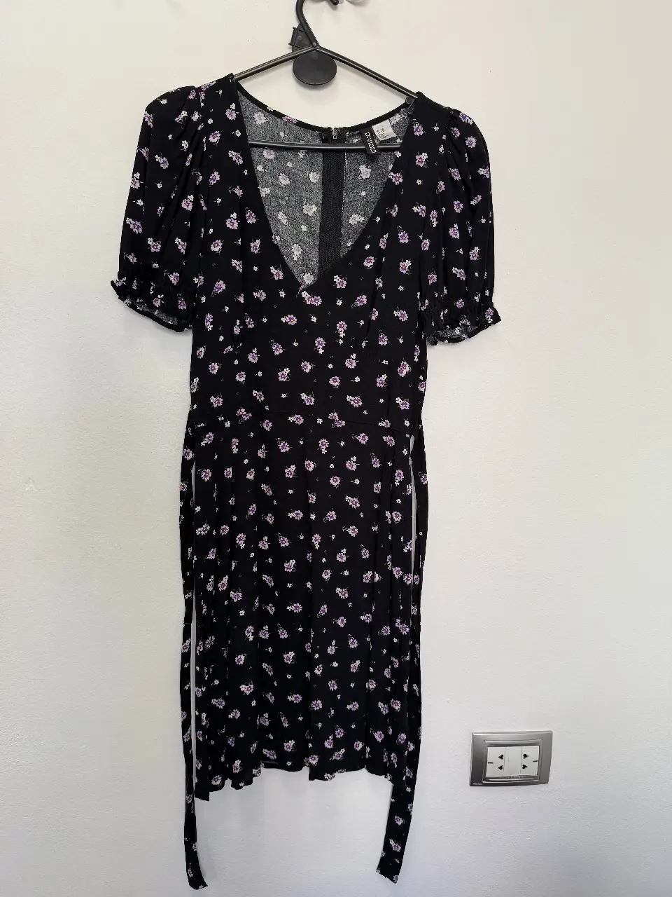 Vestido mini negro con estampado floral en tonos violetas y blancos. Corte en V, mangas cortas con volados y lazo en la cintura. Ideal para un look fresco y femenino.