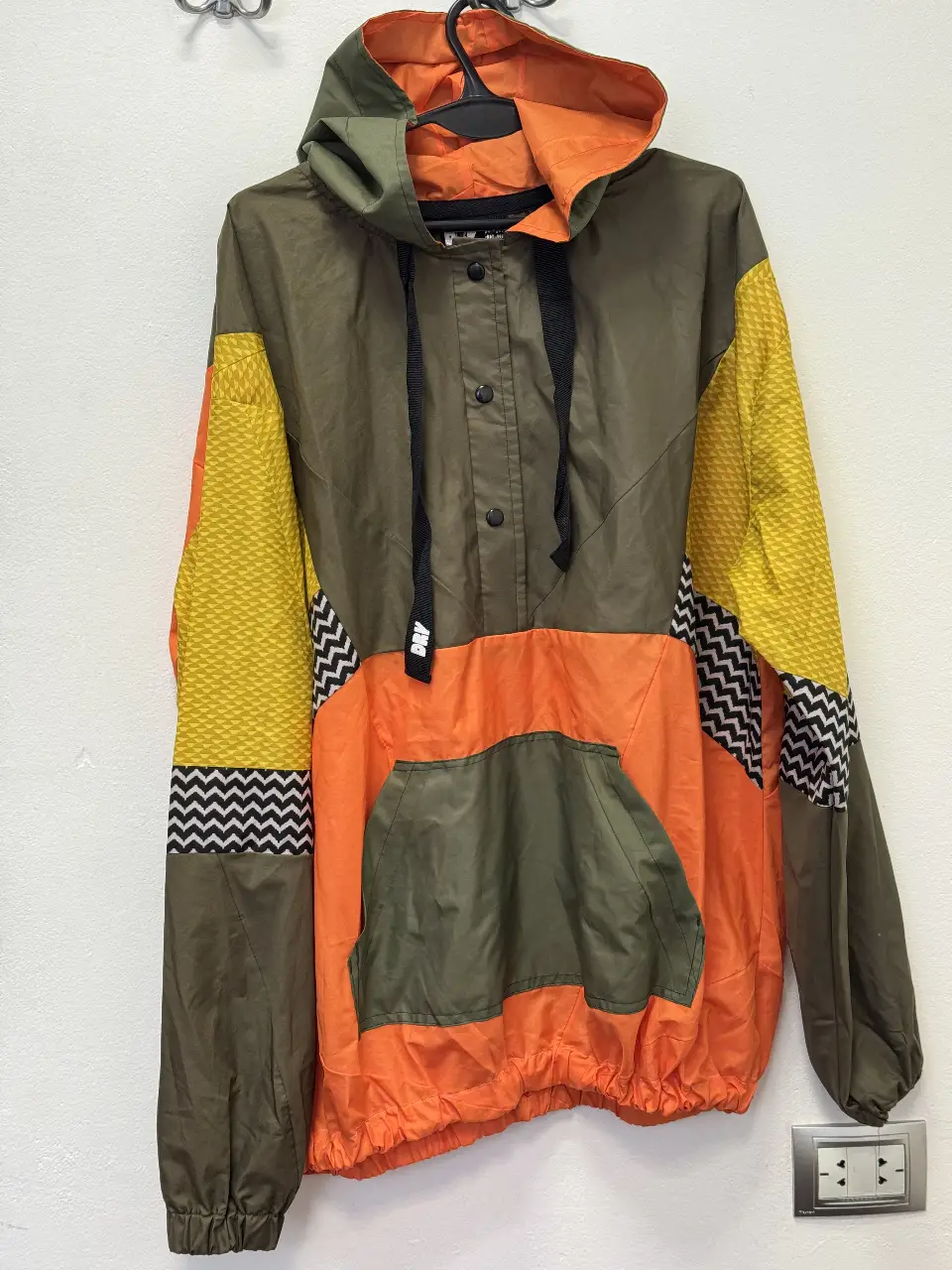 Campera rompevientos con capucha, combinada en colores verde oliva, naranja y amarillo. Detalles en negro y blanco. Ideal para un look deportivo y canchero.