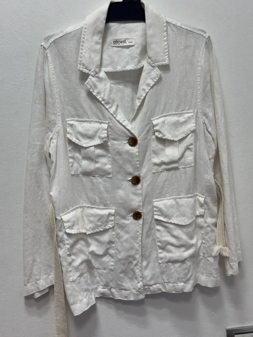 Chaqueta blazer blanca de lino, con cuatro bolsillos frontales y cinturón para ajustar la cintura. Ideal para un look casual y fresco.