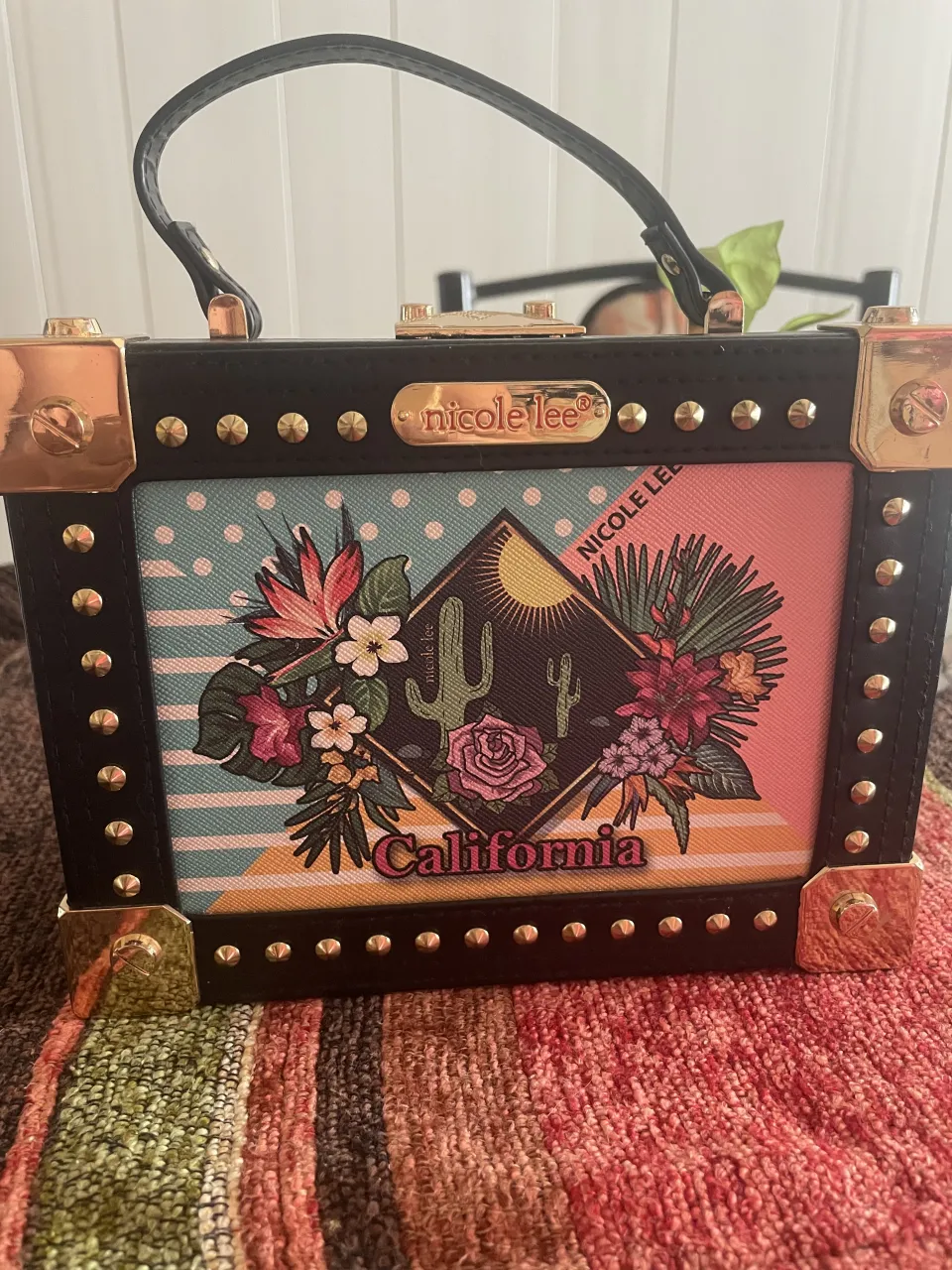 Bolso tipo baúl de Nicole Lee, con estampa floral y de cactus inspirada en California. Detalle de tachas doradas y herrajes metálicos. Ideal para darle un toque divertido y original a tu look.