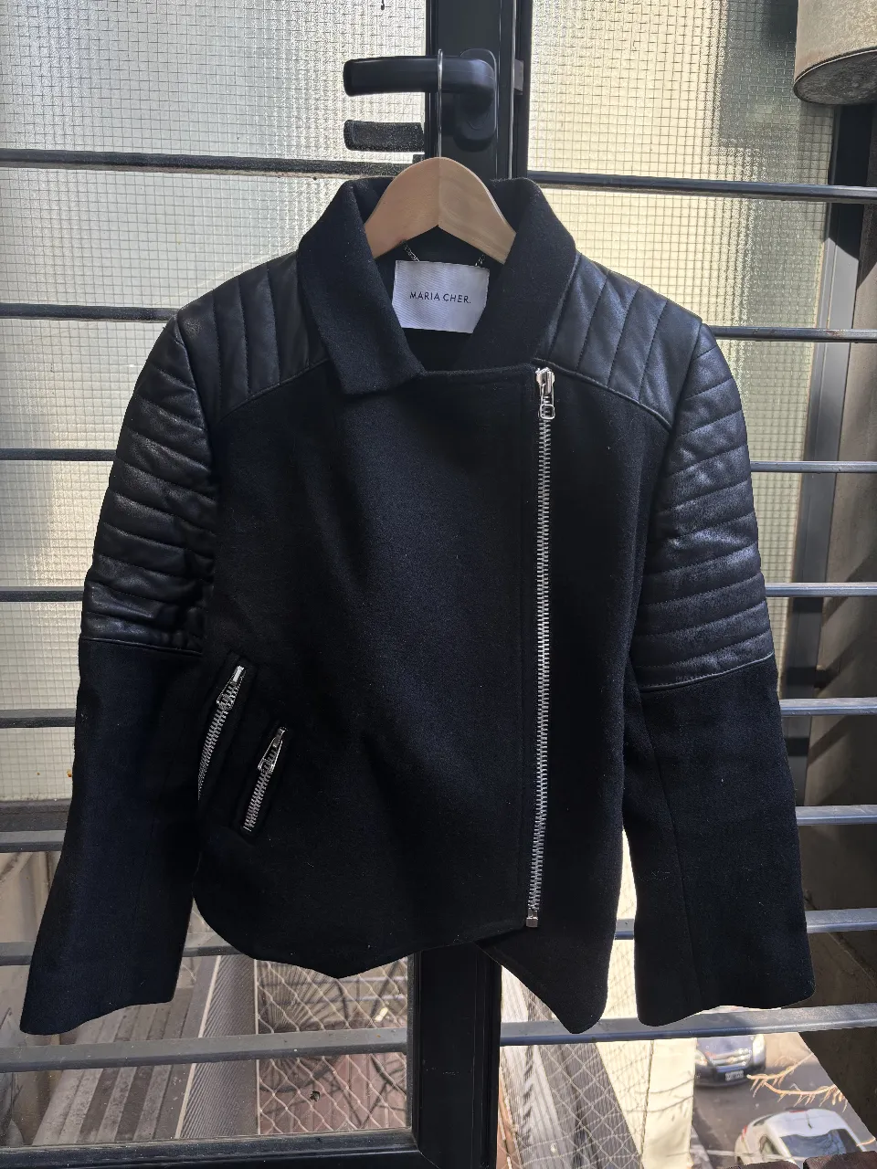 Chaqueta estilo biker para mujer, confeccionada en paño negro con detalles en cuero sintético en hombros y mangas. Diseño asimétrico con cierre y bolsillos con cremallera. Ideal para un look moderno y audaz.