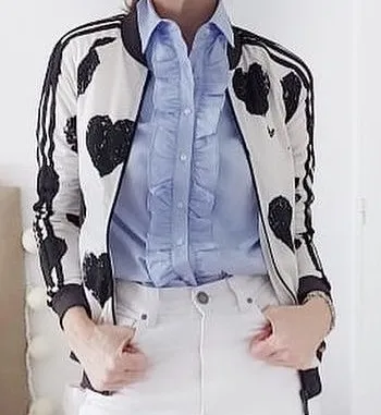 Campera bomber de Adidas con estampa de corazones negros sobre fondo blanco. Tiene cierre al frente y bolsillos laterales con cierre. Las mangas y el cuello son de ribb negro con detalles de franjas blancas.