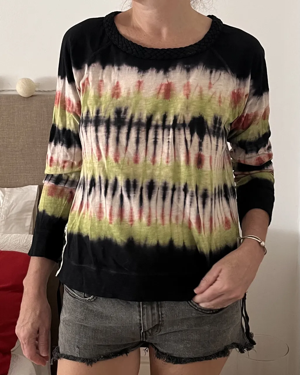 Remerabuzo manga larga con estampa tie-dye en tonos verdes, rojos y negros. Posee detalles de trenzado en el cuello y cordones en los laterales inferiores. Ideal para un look casual y relajado.