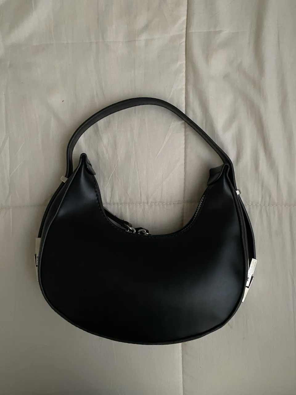 Cartera tipo baguette de color negro, con detalles plateados en los extremos y cierre con cremallera. 