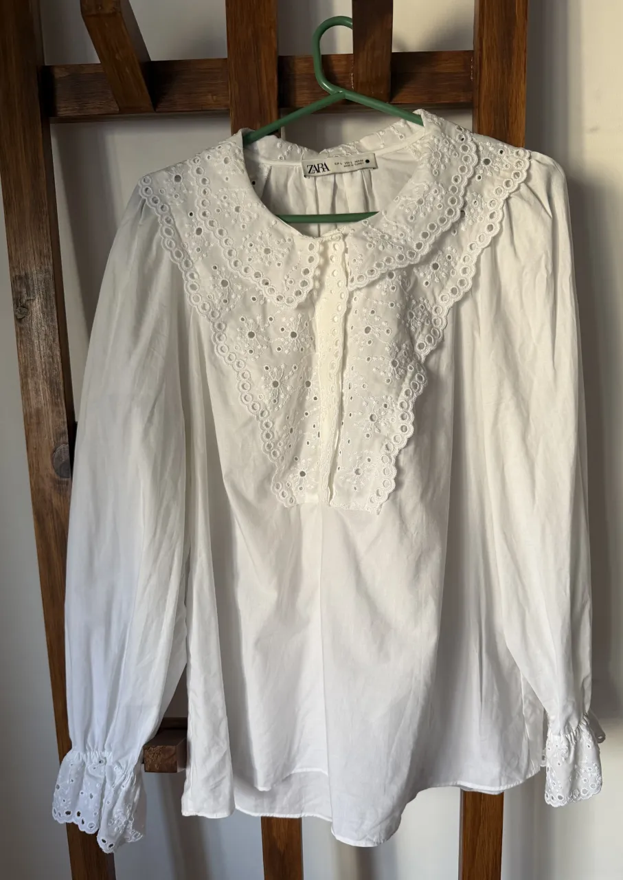 Blusa blanca de Zara con detalles de bordado y puntilla en cuello y mangas. Cuello redondo con escote en V y botones al frente. Mangas largas con puños elastizados y volado. Perfecta para un look romántico y delicado.