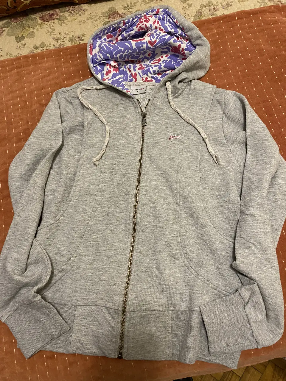Buzo con cierre gris de la marca Speedo, con capucha forrada con estampado floral en tonos violetas y rojos. Ideal para un look casual y deportivo.