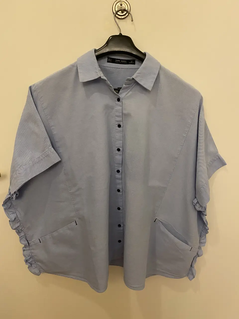 Camisa celeste de Zara Basic, con mangas cortas y detalles de volados en los laterales. Tiene dos bolsillos delanteros y cierre con botones.