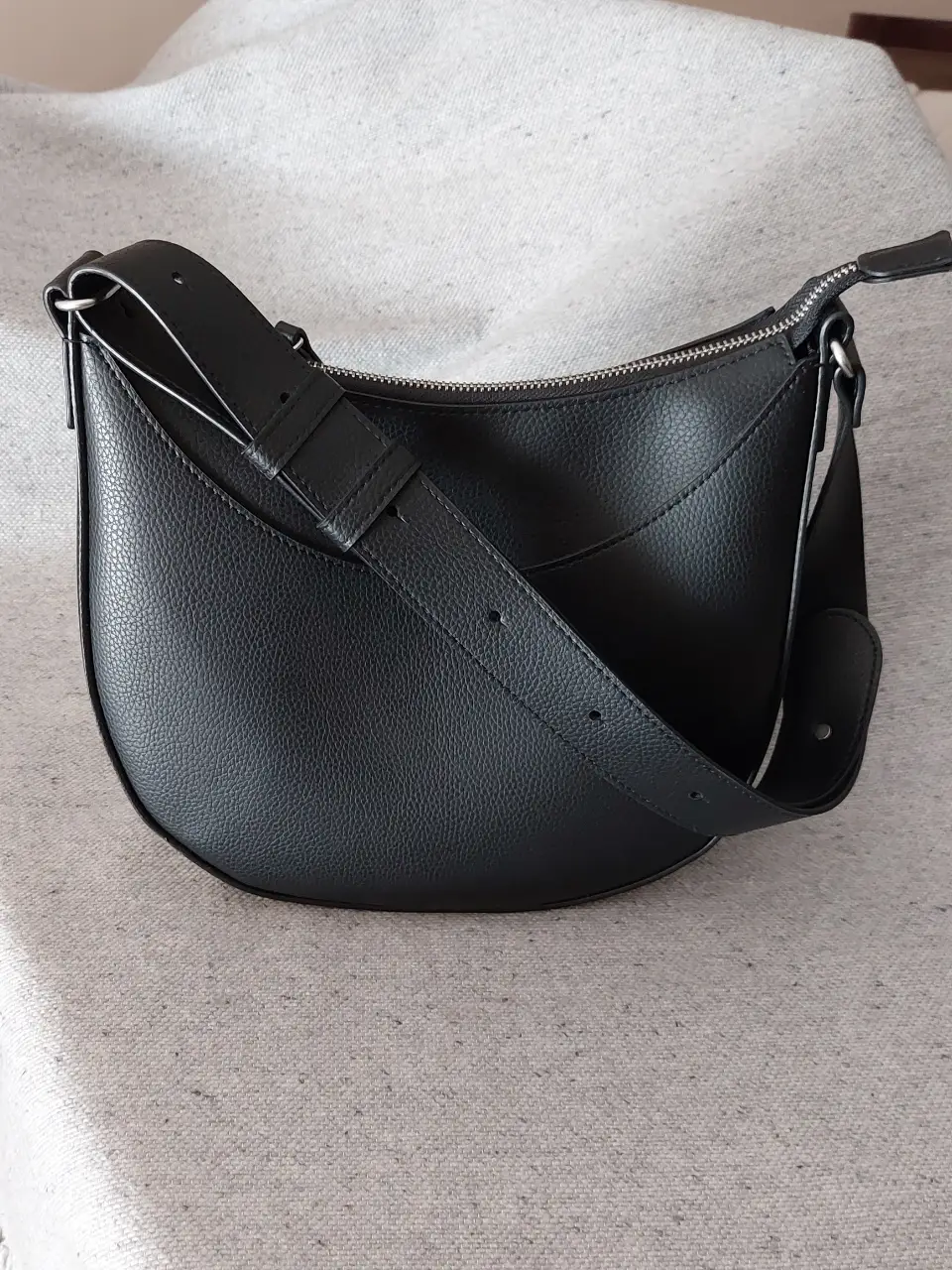 Cartera Uniqlo bandolera de cuero sintético negro, con forma de media luna. Cierre superior. Forrada en tela, con dos bolsillos internos. Correa regulable. Medidas: 21 alto x 25 ancho x 9 prof.