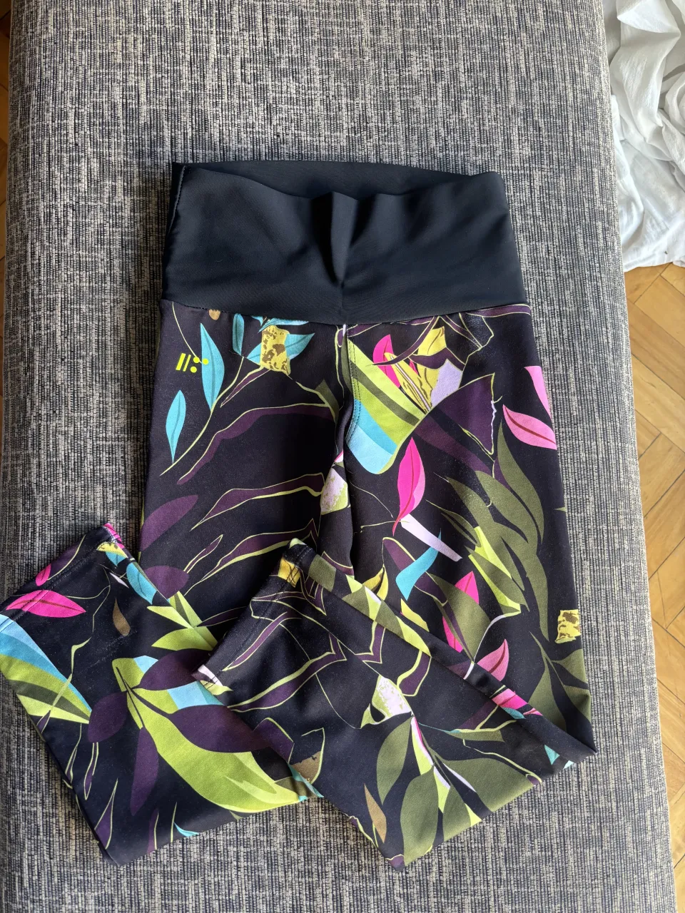 Calzas deportivas estampadas con motivos florales y hojas en tonos vibrantes sobre fondo negro. Cintura alta y cómoda, ideal para entrenar o para un look casual y canchero.