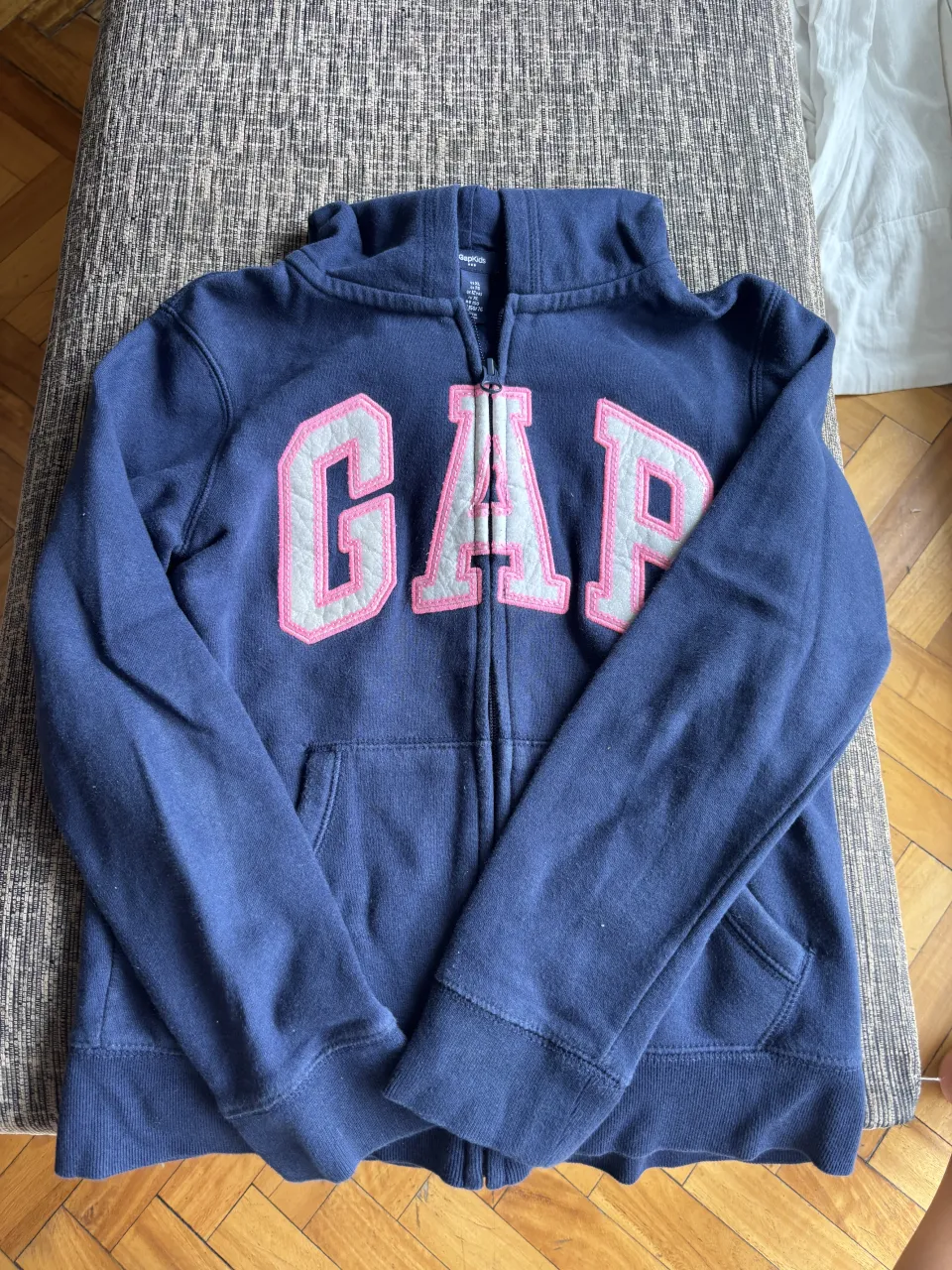 Buzo azul marino con cierre completo y capucha, de la marca Gap. Presenta el logo de la marca en grande en el frente, con letras rosas y borde gris. Tiene bolsillos tipo canguro.