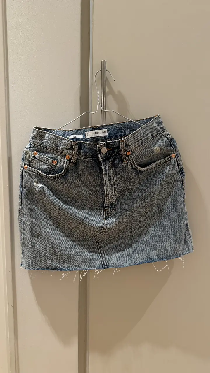 Minifalda de jean azul claro, tiro medio, con detalles de roturas y un corte desflecado en el borde inferior. Ideal para un look casual y moderno.