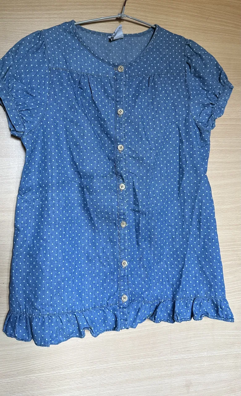 Camisa de jean azul con estampa de lunares blancos. Tiene cuello redondo, botones al frente y un detalle de volado en la parte inferior. Mangas cortas con elástico.