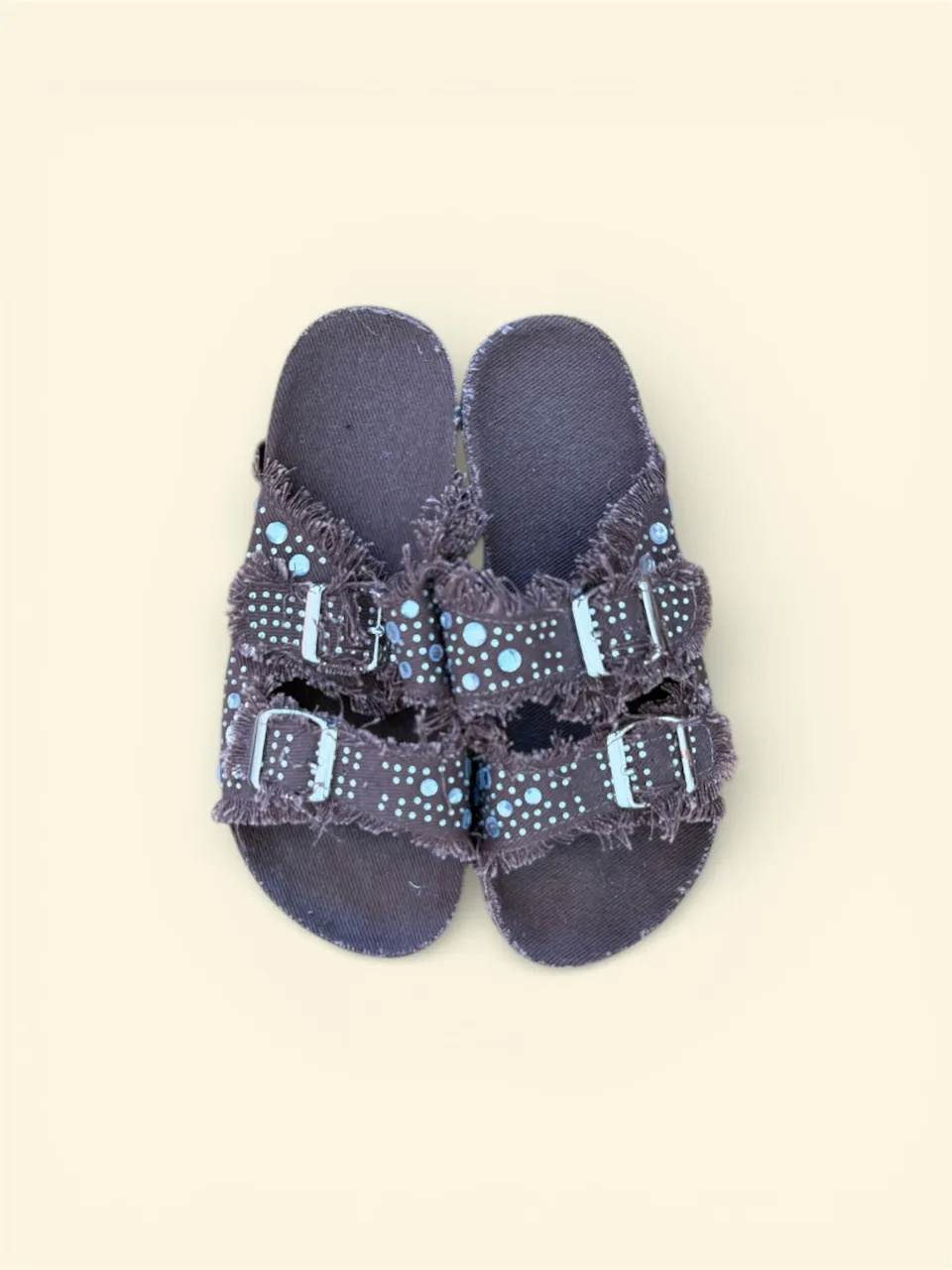 Sandalias tipo ojota de color marrón oscuro con dos tiras frontales. Las tiras tienen un borde deshilachado y están adornadas con pequeños círculos de color celeste. Cada tira tiene una hebilla metálica para ajustar.