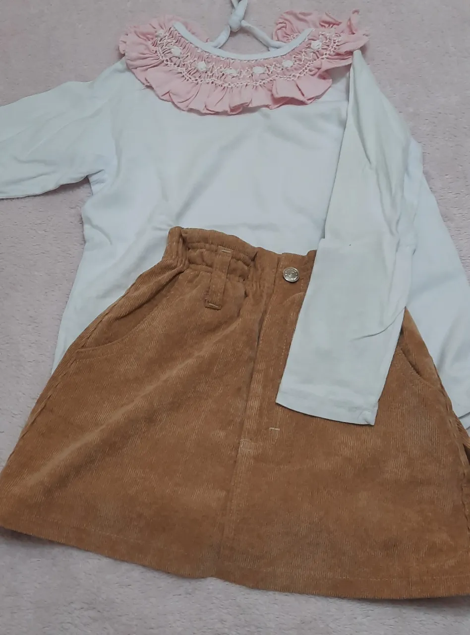 Conjunto de remera blanca de manga larga con cuello volado rosa y pollera de pana marrón con cintura elastizada y bolsillos.