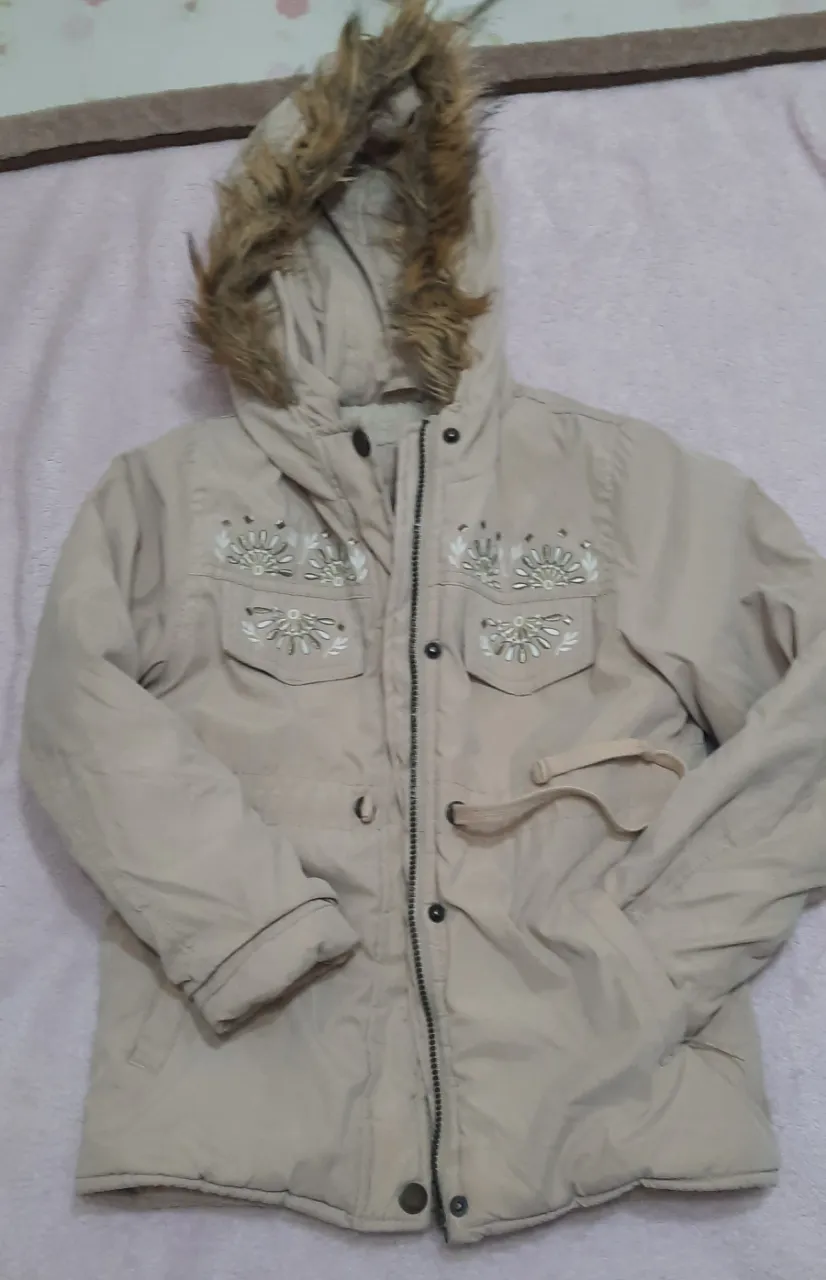 Campera beige con capucha forrada en piel sintética. Posee bordados florales en la parte delantera y bolsillos con solapa. Cierre principal y cordón ajustable en la cintura.