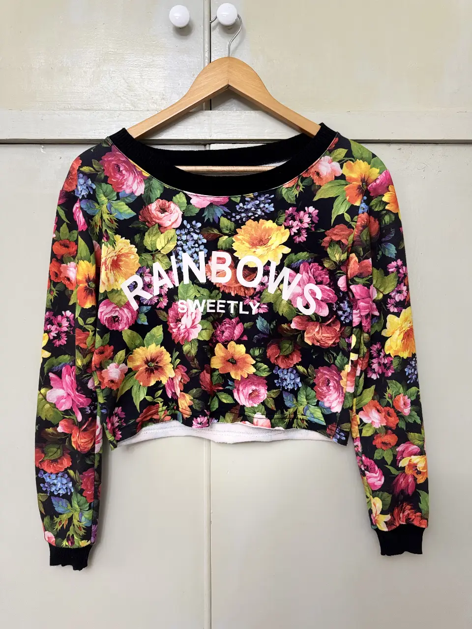 Buzo crop con estampado floral y la frase "RAINBOWS SWEETLY" en blanco. Ideal para un look casual y moderno.