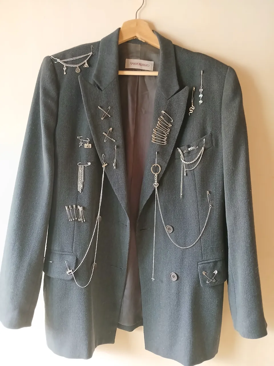 Blazer gris oscuro con detalles punk, adornado con alfileres de gancho y cadenas plateadas. Un toque de rebeldía para un look moderno y audaz.