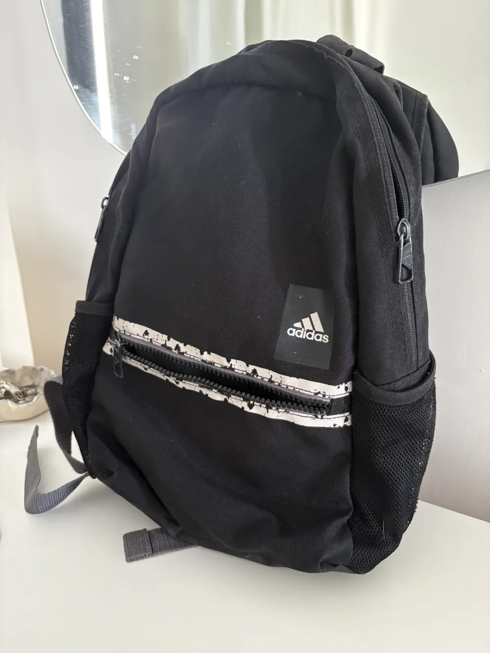 Mochila negra de tela con logo de Adidas blanco y detalles de líneas blancas y salpicaduras en el bolsillo frontal. Tiene bolsillos laterales de red y correas ajustables.
mide 35 cm de largo x 27 cm de ancho aprox. es chica ideal para el gimnasio 